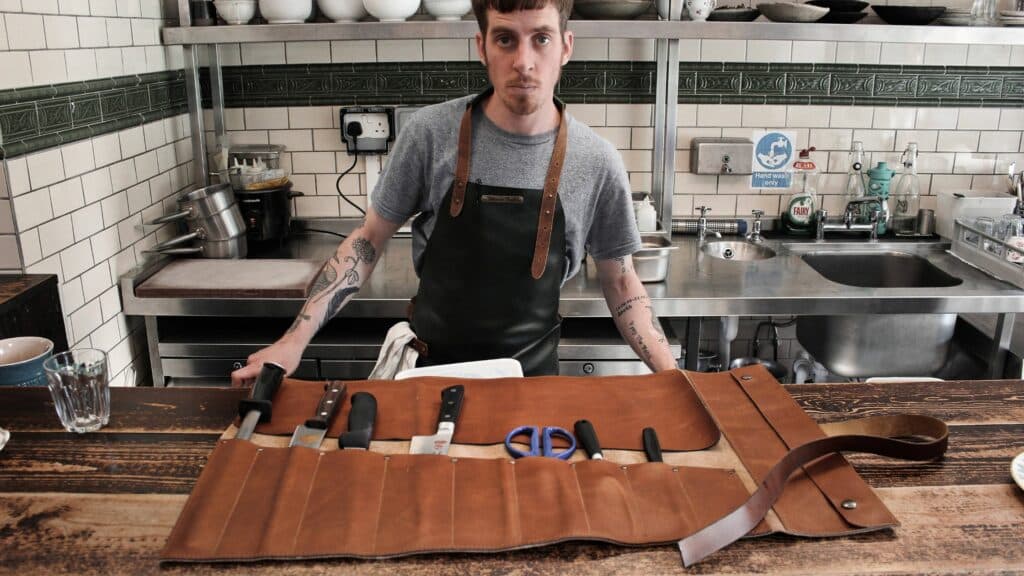 Leather apron - 5 professions that need them. Image of a man with a leather apron that holds his kitchen knife set for cooking